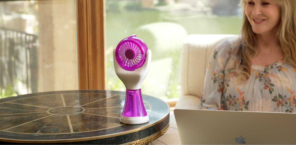 A woman sits at a table by a window using a laptop, with a small purple and white handheld fan placed on the table beside her.