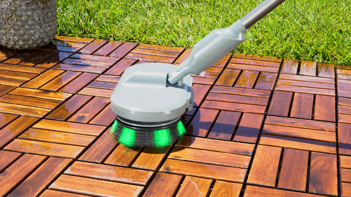 A close-up of a cleaning brush with a gray handle and green bristles, cleaning a brown wooden deck outdoors next to green grass.