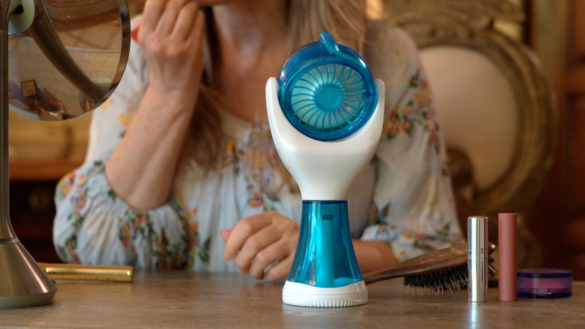 A handheld electric fan with a blue and white design sits on a wooden table in a vanity setting, with a woman applying makeup in the background.