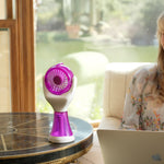 A woman sits at a table by a window using a laptop, with a small purple and white handheld fan placed on the table beside her.