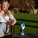 A woman sits in a garden chair outdoors, reading a book or magazine while wearing a floral long-sleeve top and jeans. A small blue-and-white handheld device sits on a metal table beside her.