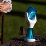 A handheld personal fan with a translucent blue and white design sits on a dark metal patio table outdoors, with a person reading in a chair blurred in the background on a green lawn.