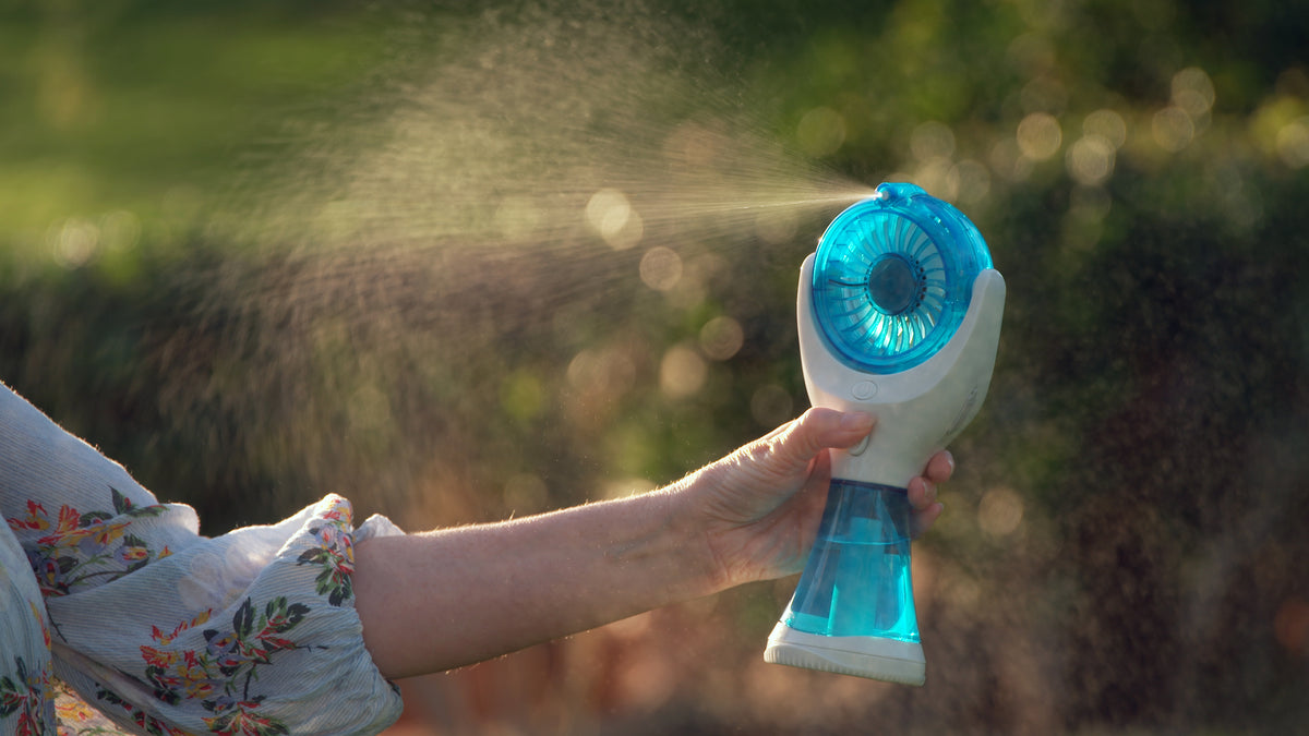 A handheld personal fan with a blue translucent face and white body, being held by a person outdoors. A fine mist is spraying from the fan, with a blurred green and brown natural background.