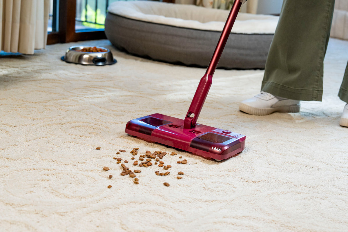 A person using a red and black cordless stick vacuum cleaner to clean a beige carpet, with scattered kibble on the floor nearby. The person is wearing green pants and white sneakers, and a pet bed and bowl are visible in the background.