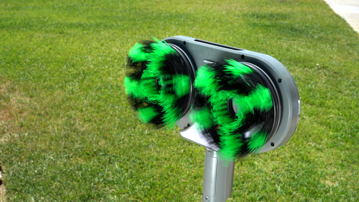 A close-up of a gray automatic coin-operated cleaning brush machine with two spinning green-and-black striped brushes, set against a background of green grass.