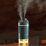 A small tabletop humidifier emitting a fine mist, shown on a reflective surface with a softly blurred indoor background.