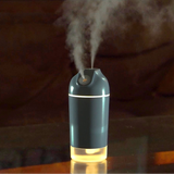 A small tabletop humidifier emitting a fine mist, shown on a reflective surface with a softly blurred indoor background.