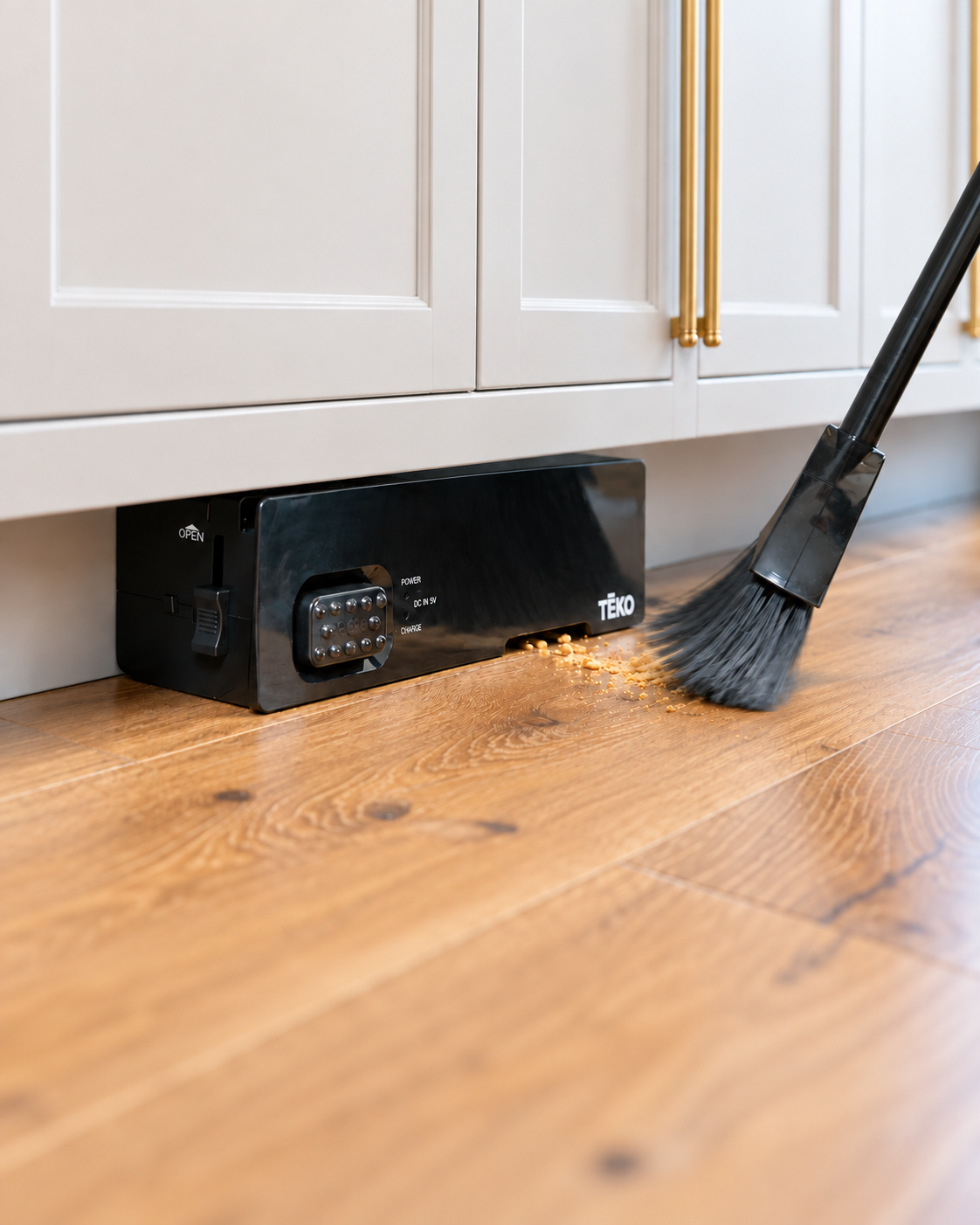 A black Teko vacuum placed under white baseboards, with a broom sweeping debris toward it on a hardwood floor.