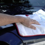 A close-up of a person's hand wiping down the hood of a black car with a white microfiber cloth that has a red border.