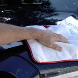 A close-up of a person's hand wiping down the hood of a black car with a white microfiber cloth that has a red border.