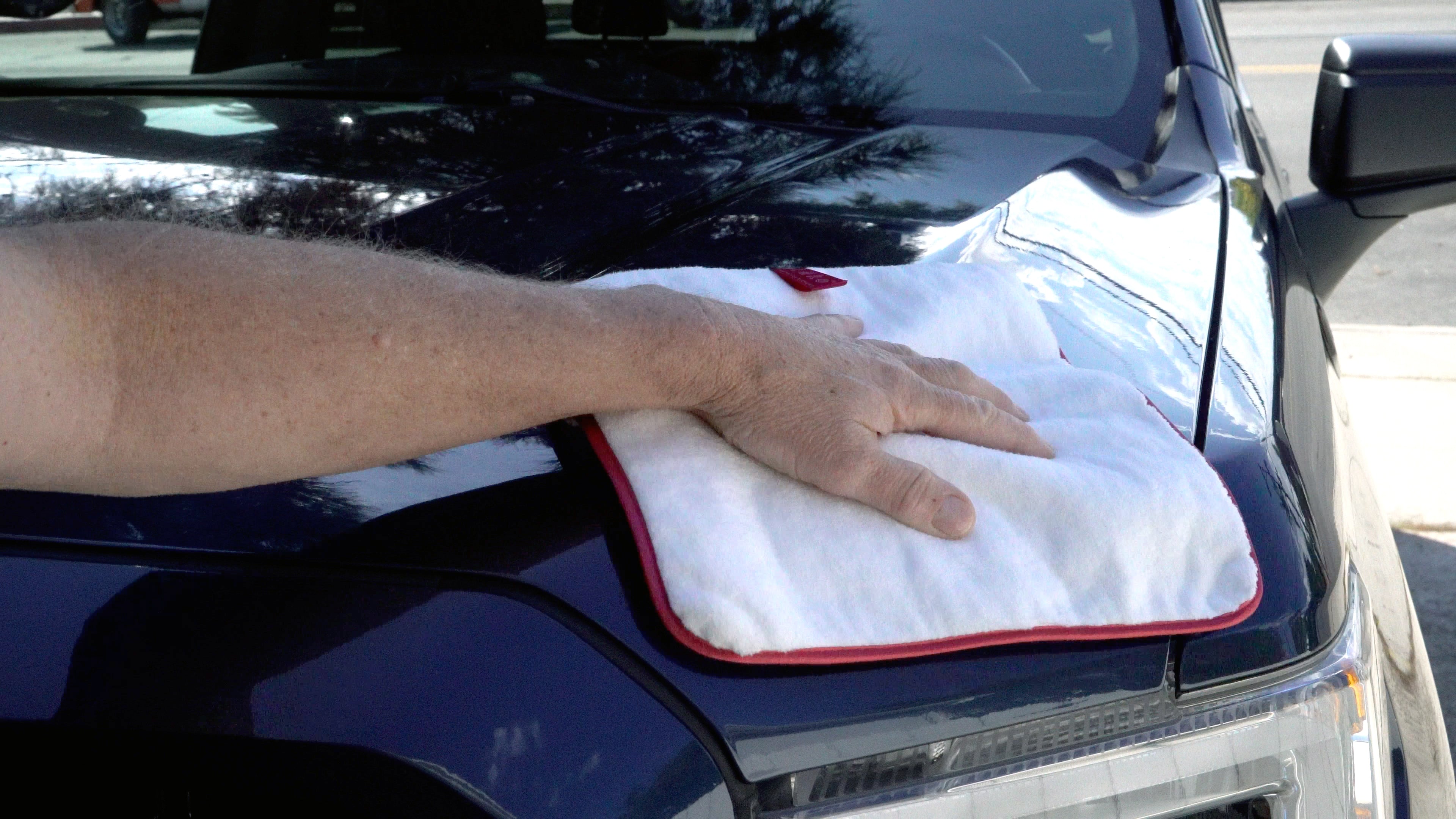 A close-up of a person's hand wiping down the hood of a black car with a white microfiber cloth that has a red border.