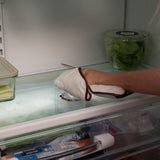 A person wearing a white oven mitt with brown trim reaches inside a refrigerator to grab a glass container on the shelf.