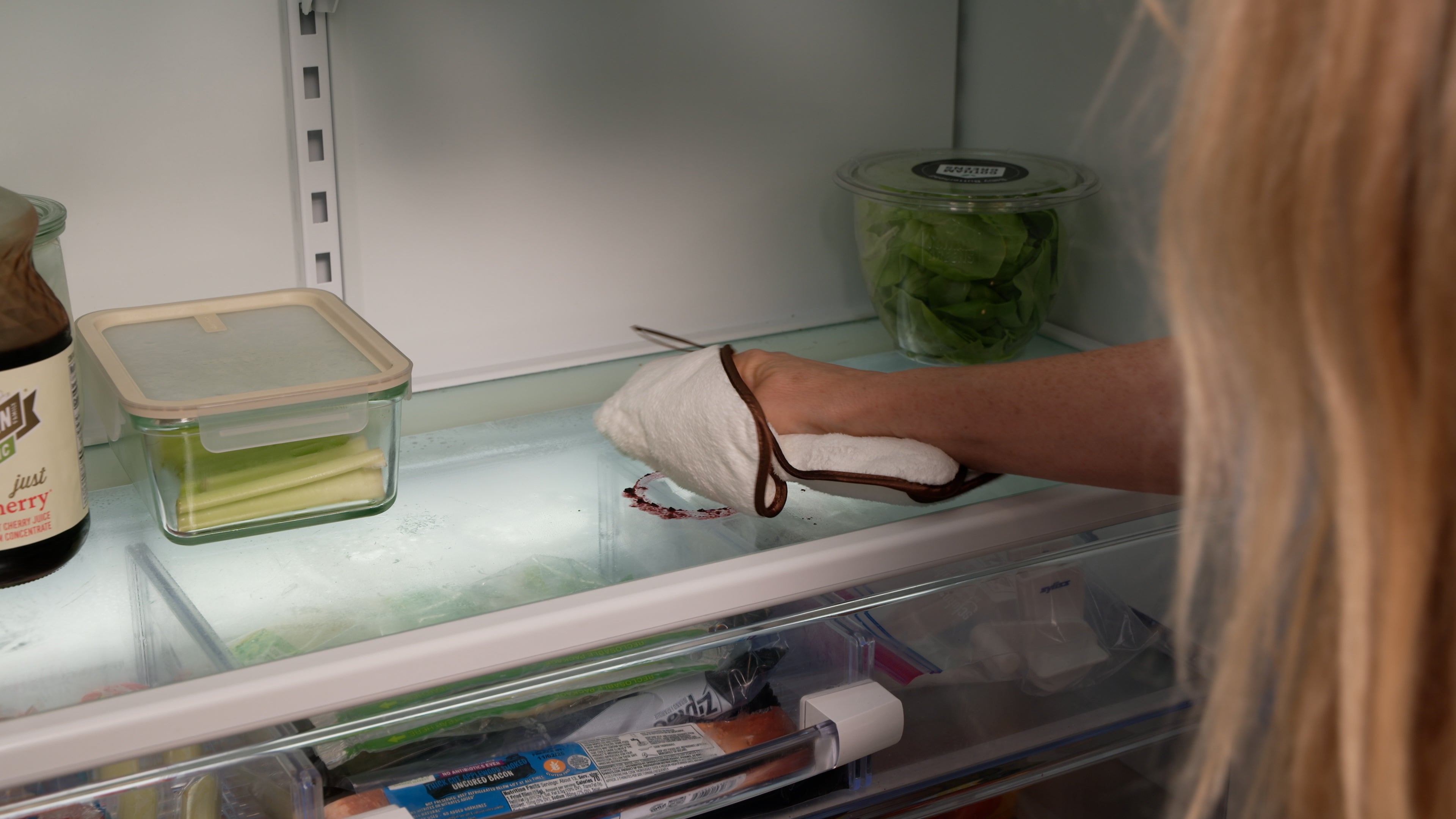 A person wearing a white oven mitt with brown trim reaches inside a refrigerator to grab a glass container on the shelf.