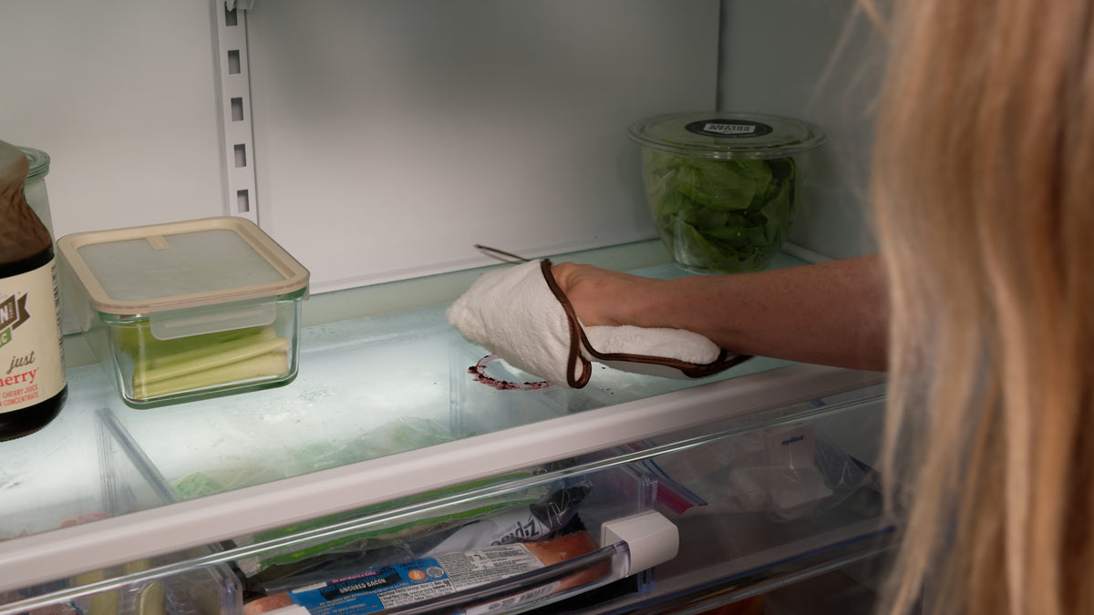 A person wearing a white oven mitt with brown trim reaches inside a refrigerator to grab a glass container on the shelf.
