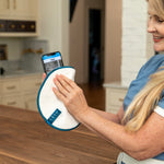 A woman in a blue scrub top holds a white circular cleaning pad with a blue edge and a smartphone showing a tracking app on a wooden table in a kitchen setting.