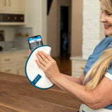 A woman in a blue scrub top holds a white circular cleaning pad with a blue edge and a smartphone showing a tracking app on a wooden table in a kitchen setting.