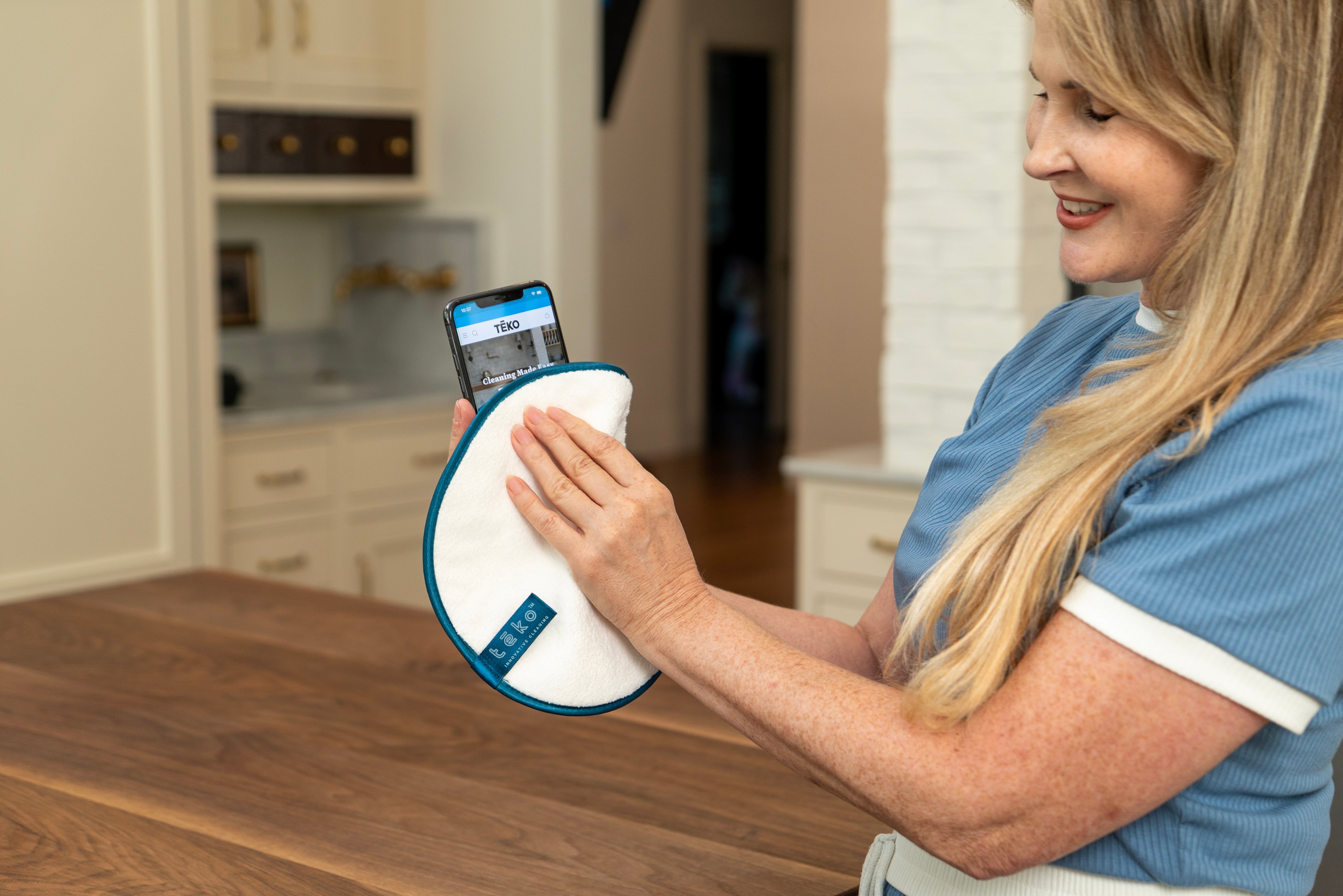 A woman in a blue scrub top holds a white circular cleaning pad with a blue edge and a smartphone showing a tracking app on a wooden table in a kitchen setting.
