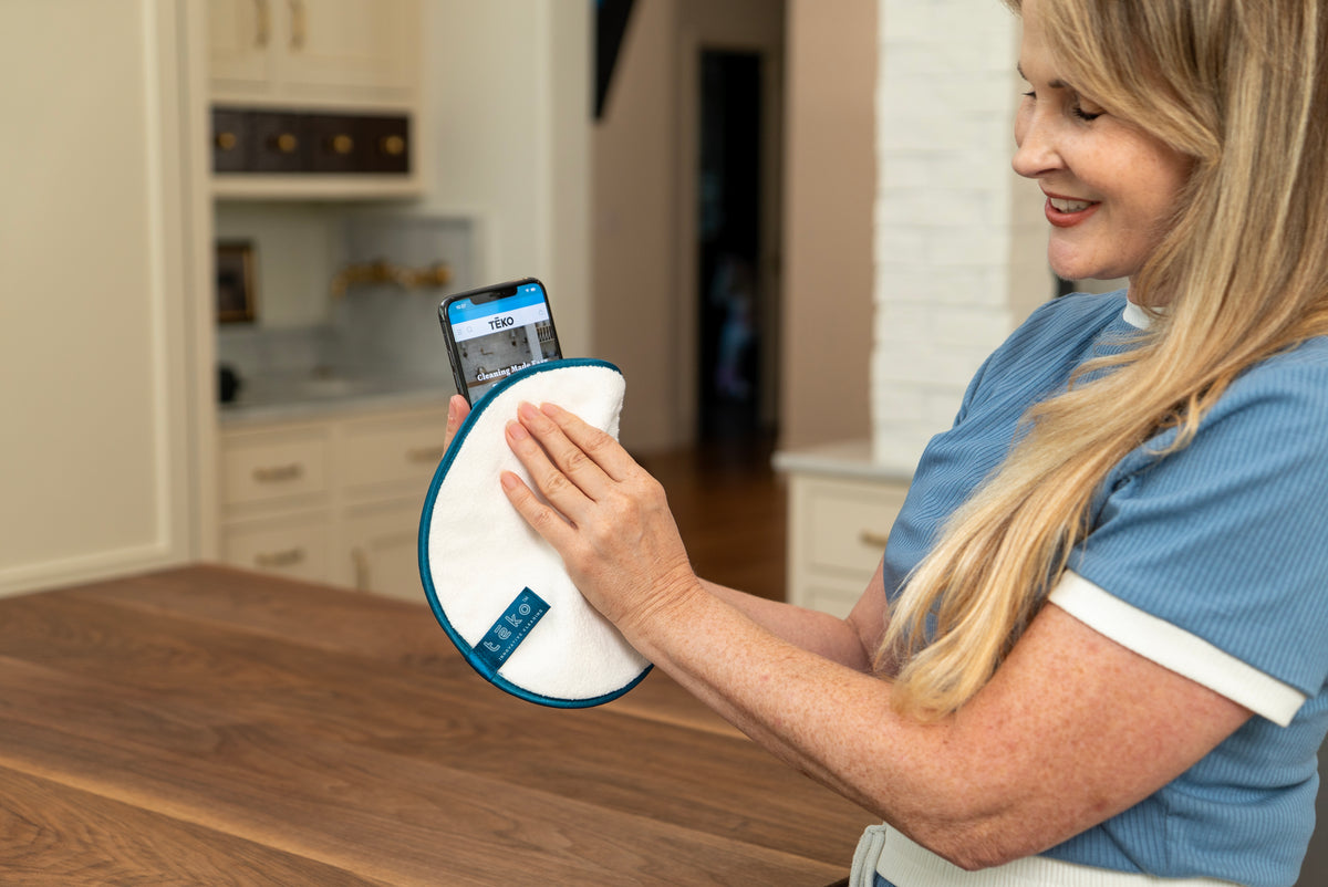 A woman in a blue scrub top holds a white circular cleaning pad with a blue edge and a smartphone showing a tracking app on a wooden table in a kitchen setting.