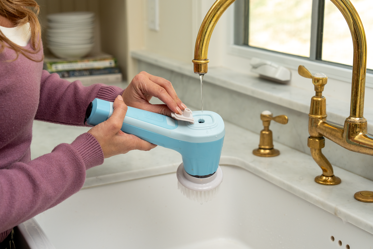 A person in a purple sweater holds a light blue and white handheld scrubber under running water in a kitchen sink with a gold faucet.