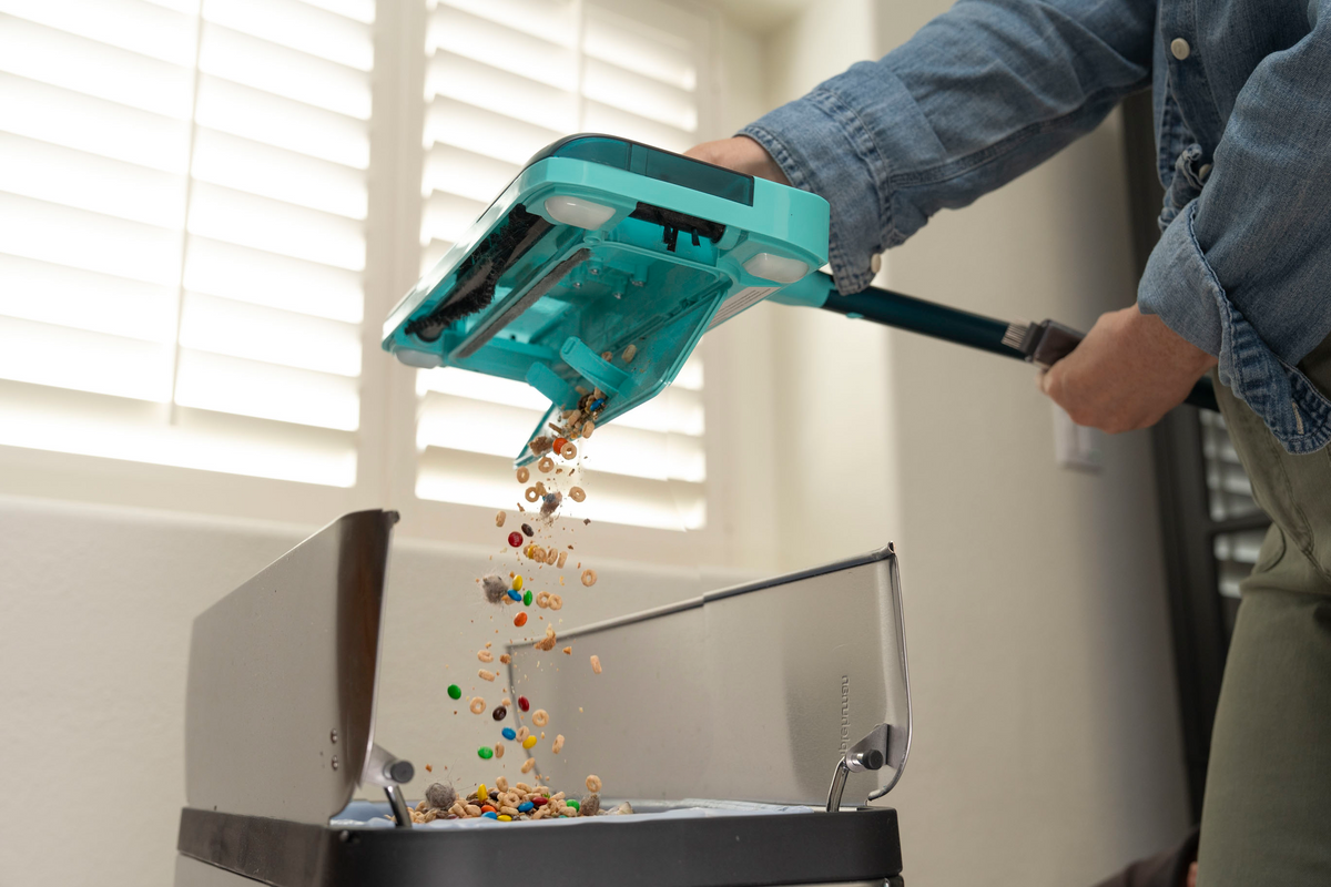 A person in a denim shirt uses a teal floor sweeper to empty colorful candy-like debris into a waste basket, shown in a home setting.