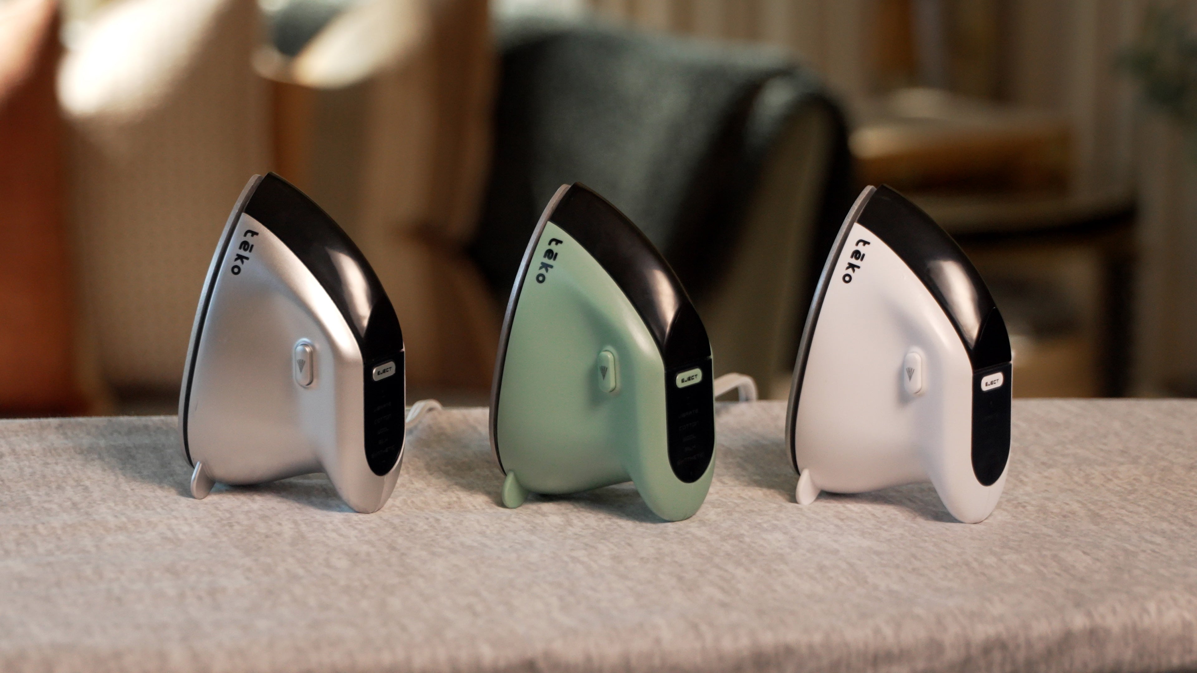 Three compact hair styling tools with curved ergonomic designs are lined up on a textured gray surface. From left to right, the colors are metallic silver, matte sage green, and glossy white, each featuring a black accent panel and a small white button.