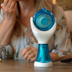A handheld electric fan with a blue and white design sits on a wooden table in a vanity setting, with a woman applying makeup in the background.