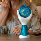 A handheld electric fan with a blue and white design sits on a wooden table in a vanity setting, with a woman applying makeup in the background.