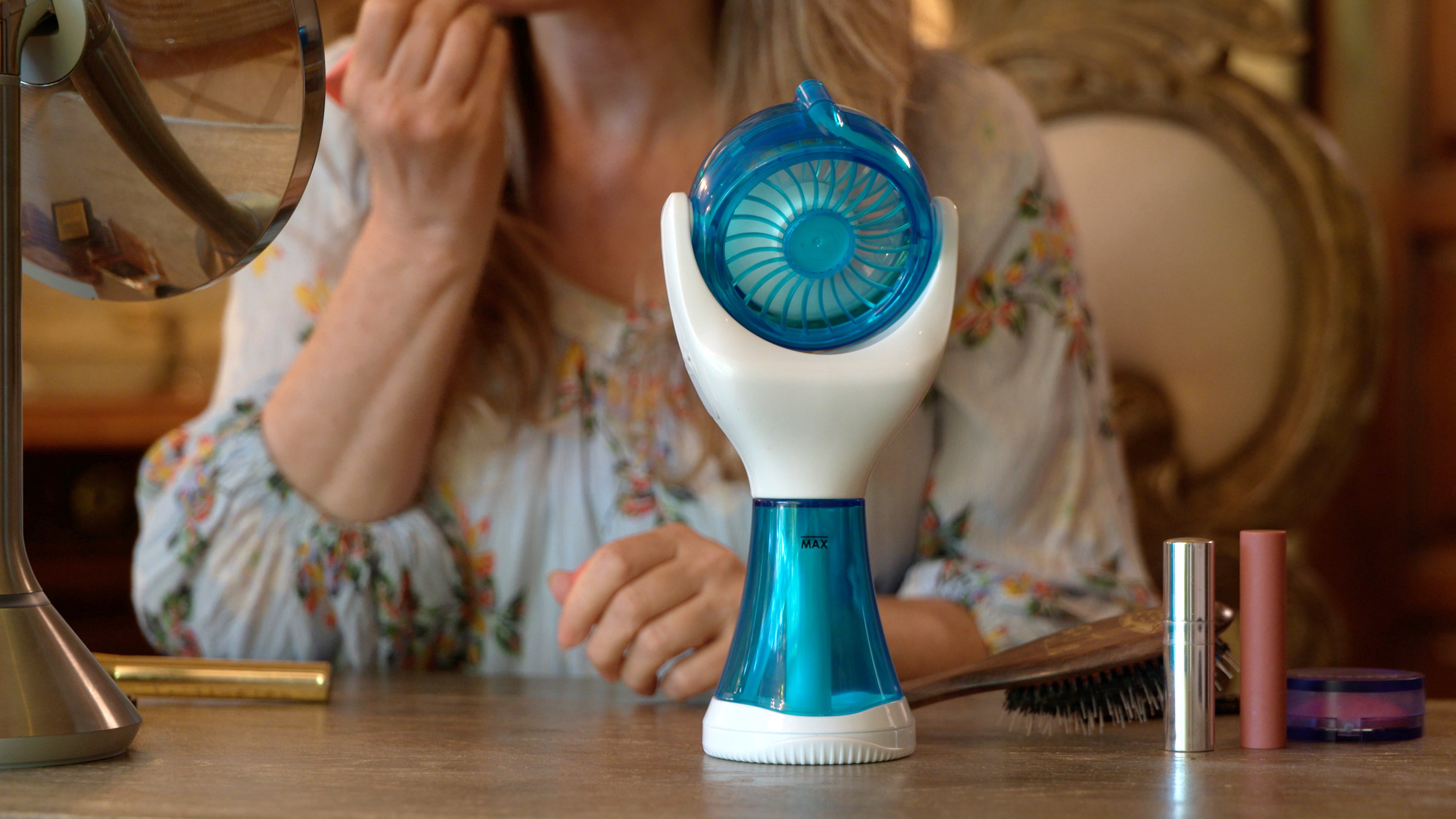 A handheld electric fan with a blue and white design sits on a wooden table in a vanity setting, with a woman applying makeup in the background.