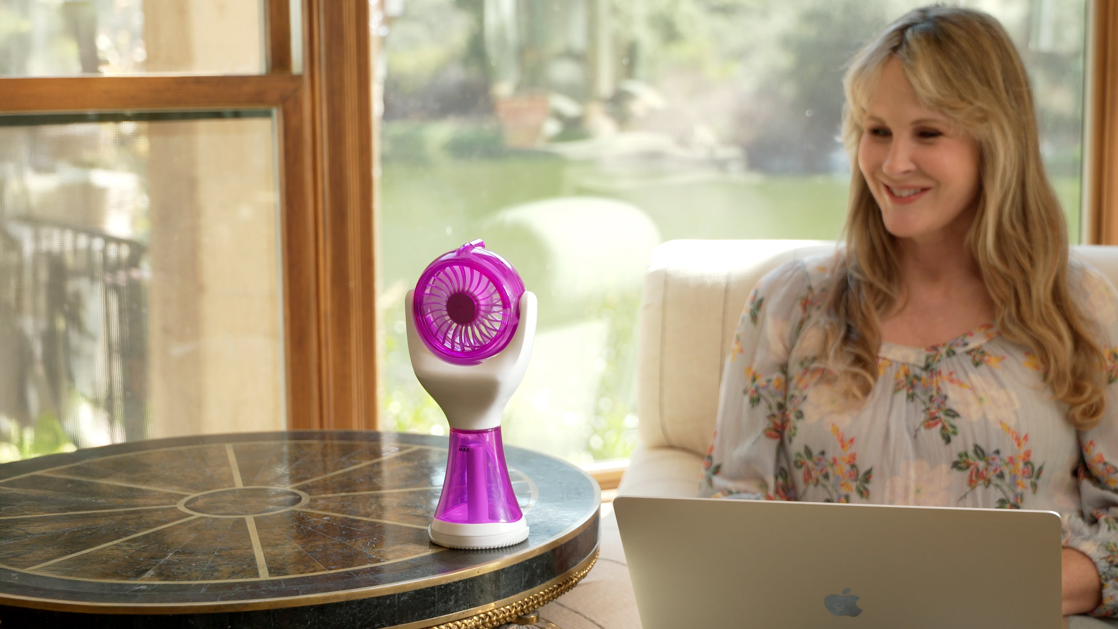 A woman sits at a table by a window using a laptop, with a small purple and white handheld fan placed on the table beside her.