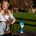 A woman sits in a garden chair outdoors, reading a book or magazine while wearing a floral long-sleeve top and jeans. A small blue-and-white handheld device sits on a metal table beside her.