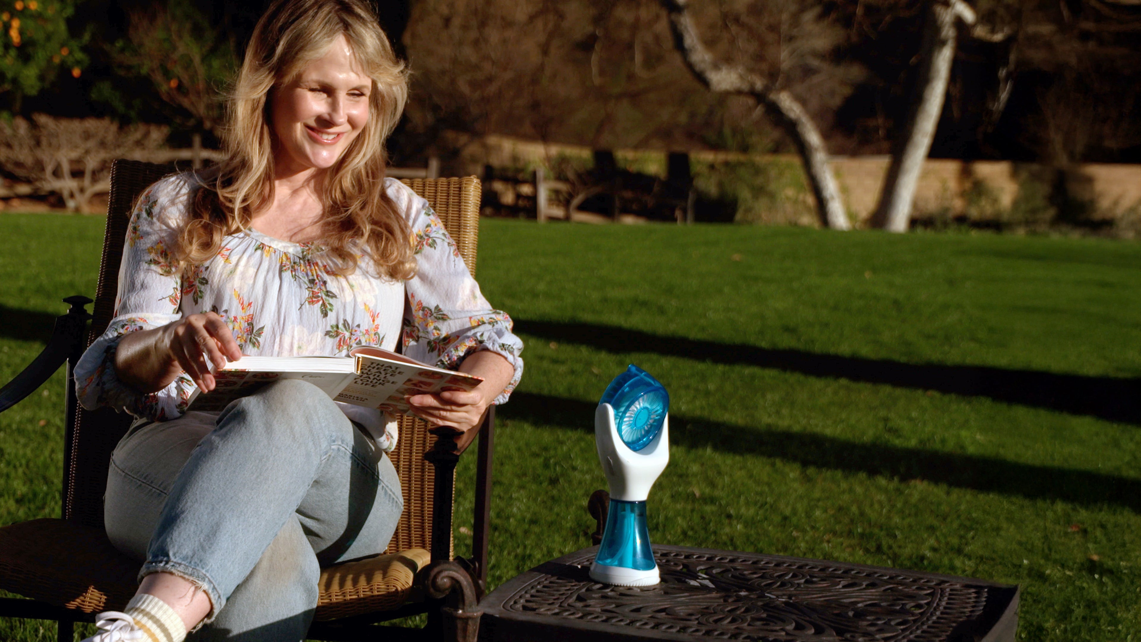 A woman sits in a garden chair outdoors, reading a book or magazine while wearing a floral long-sleeve top and jeans. A small blue-and-white handheld device sits on a metal table beside her.