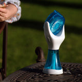 A handheld personal fan with a translucent blue and white design sits on a dark metal patio table outdoors, with a person reading in a chair blurred in the background on a green lawn.