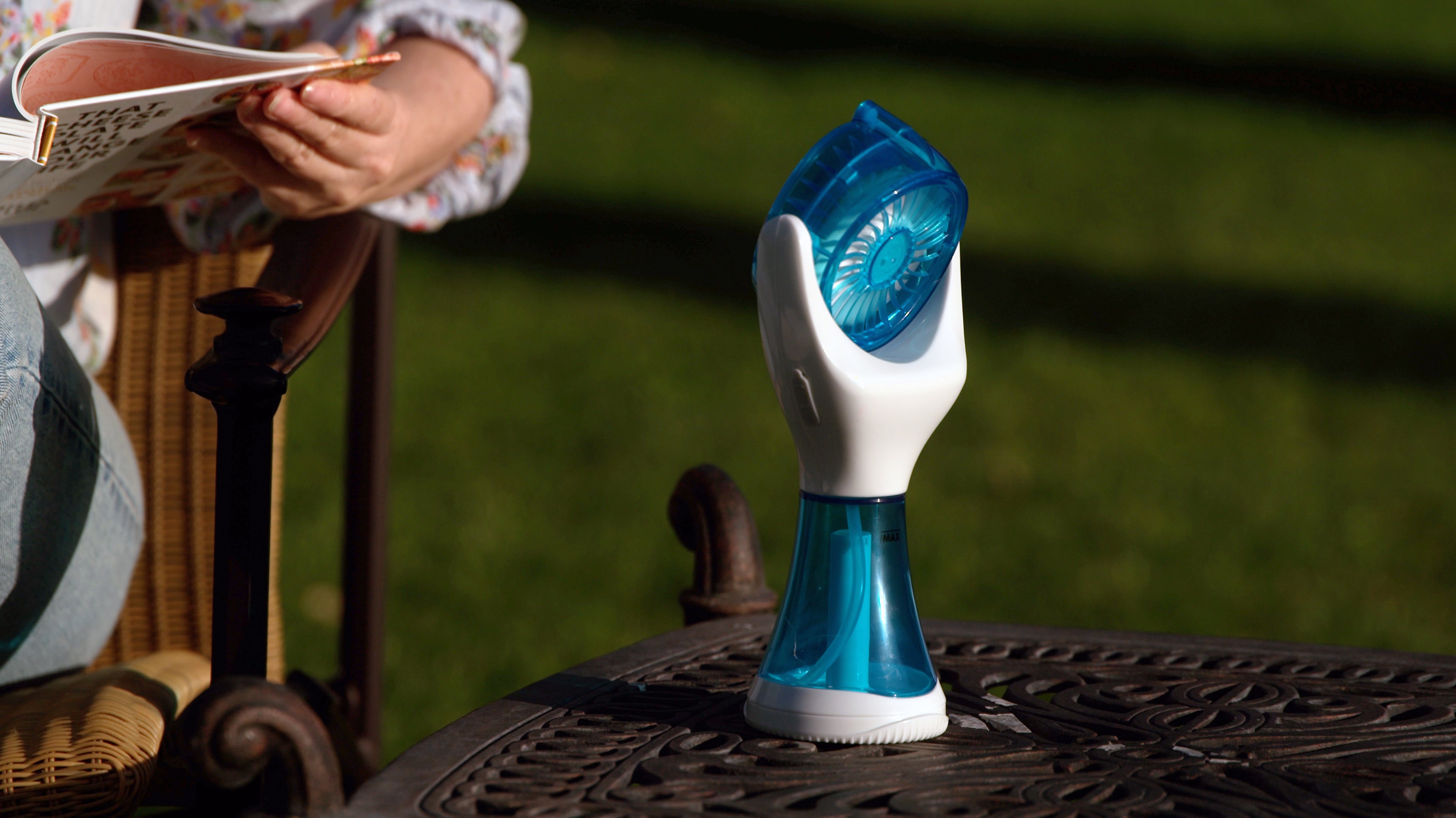 A handheld personal fan with a translucent blue and white design sits on a dark metal patio table outdoors, with a person reading in a chair blurred in the background on a green lawn.