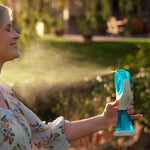 A woman outdoors in a garden holding a handheld misting spray bottle that releases a fine water spray. She is wearing a light floral blouse, and the background shows blurred greenery and warm sunlight.