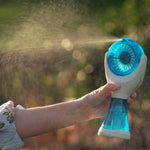 A handheld personal fan with a blue translucent face and white body, being held by a person outdoors. A fine mist is spraying from the fan, with a blurred green and brown natural background.