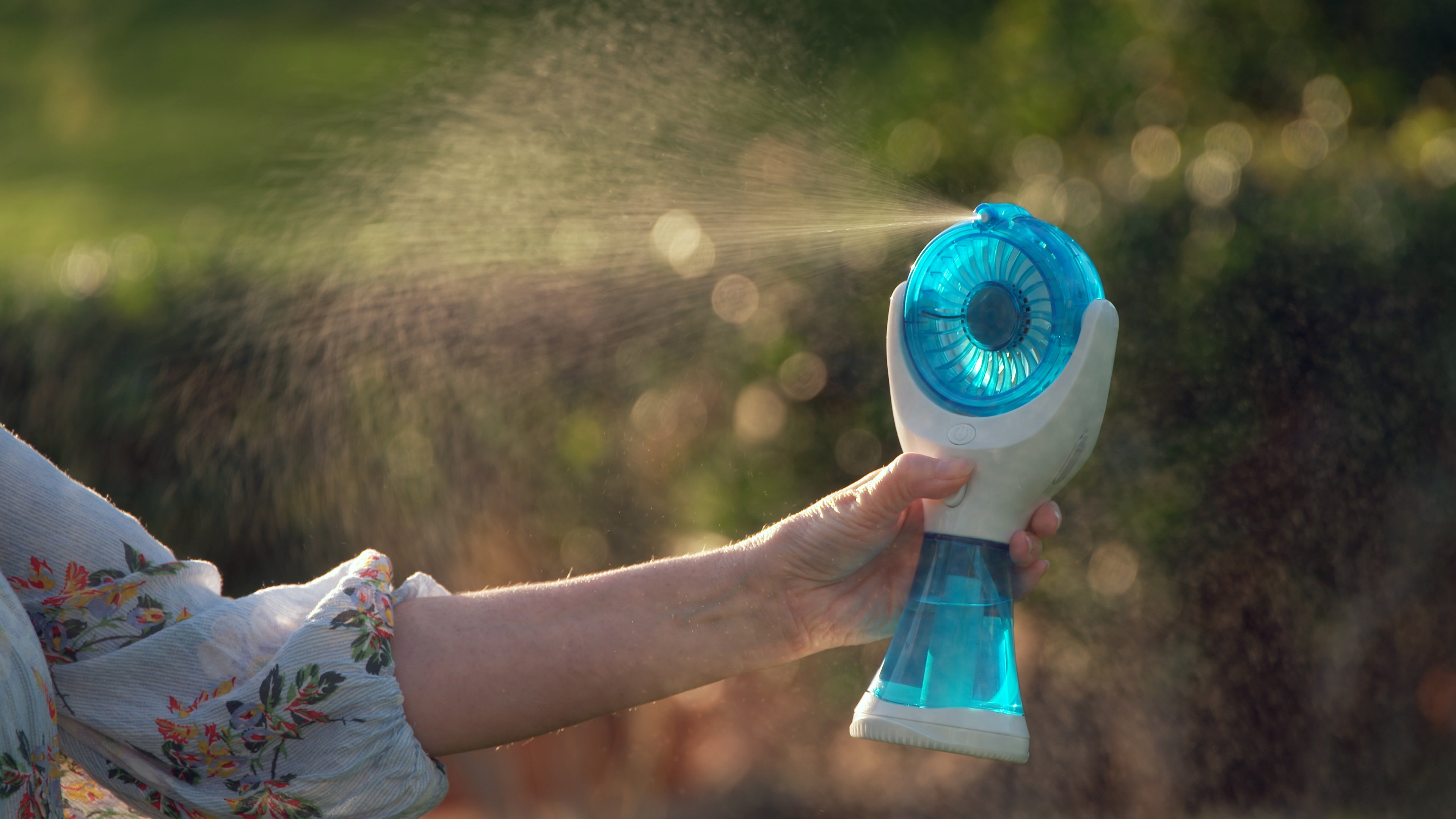 A handheld personal fan with a blue translucent face and white body, being held by a person outdoors. A fine mist is spraying from the fan, with a blurred green and brown natural background.