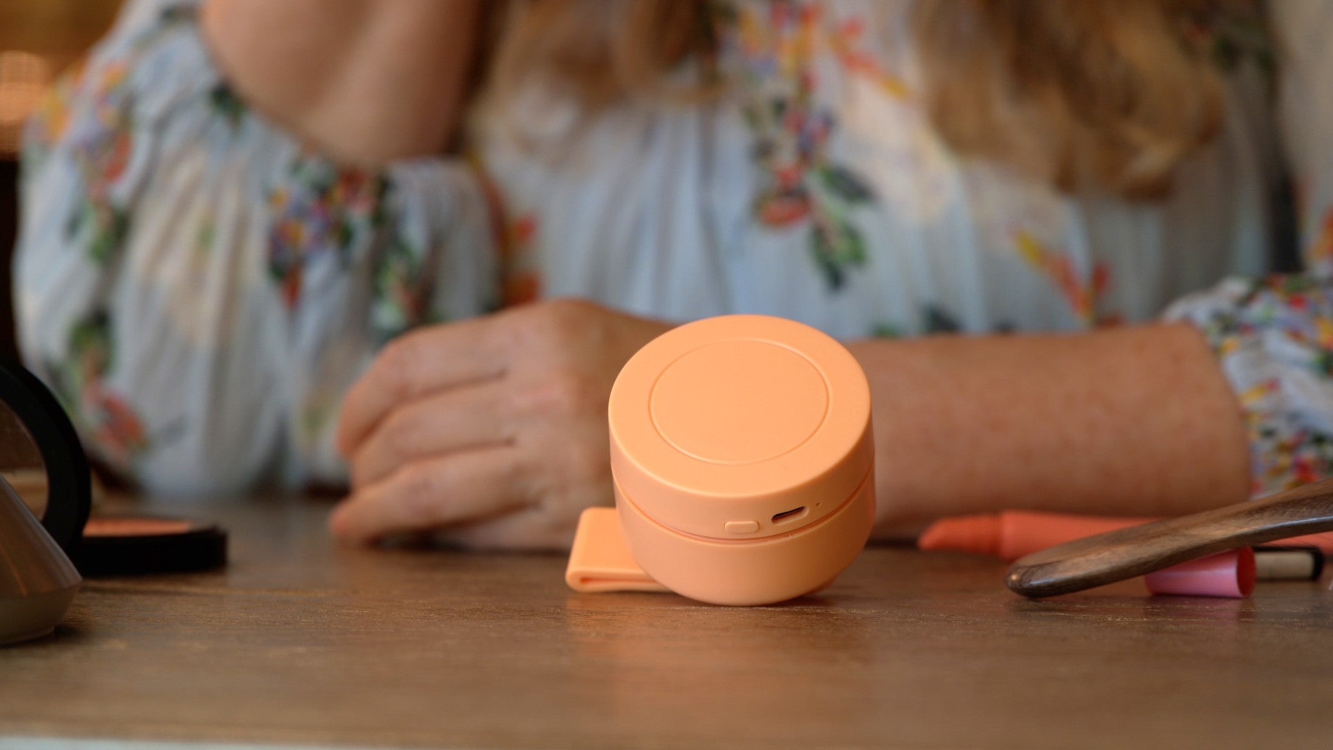 A small round peach-colored portable power bank or charging case sits on a wooden table, with a person in a floral blouse blurred in the background.