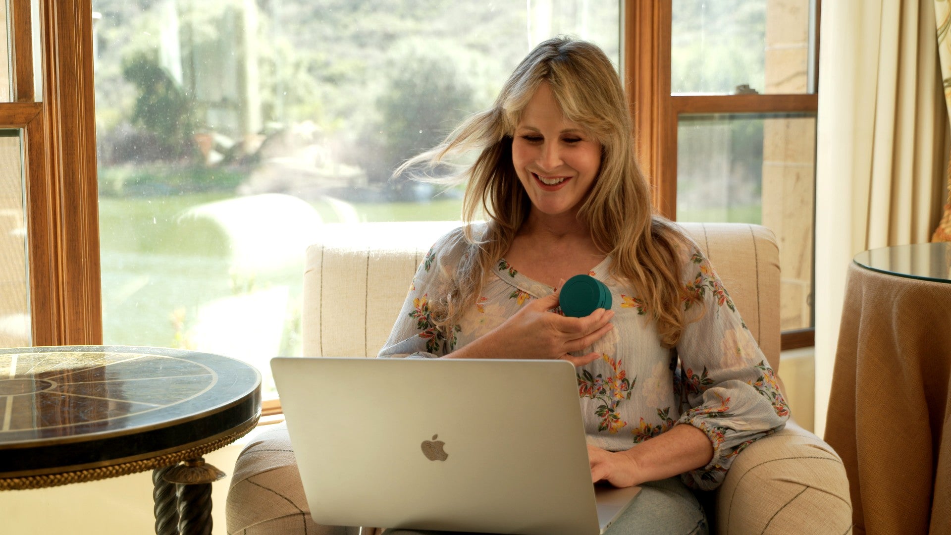 A woman sitting in a beige armchair in front of large windows, holding a small teal circular object and looking at a silver laptop. She is wearing a light floral blouse.