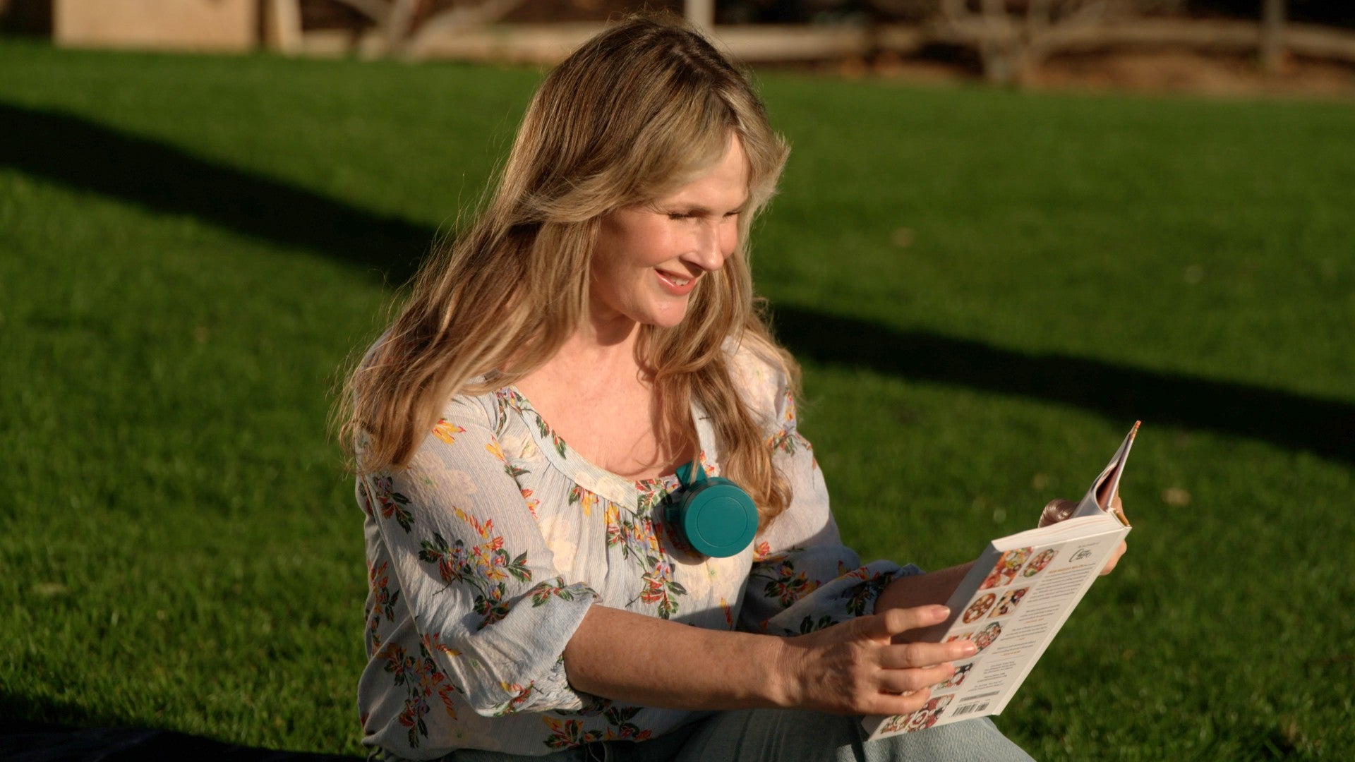 A woman sitting on a green lawn outdoors, smiling while reading a magazine. She is wearing a light floral blouse and light blue jeans, with a teal circular pendant necklace.