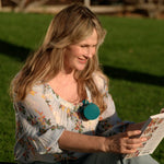 A woman sitting on a green lawn outdoors, smiling while reading a magazine. She is wearing a light floral blouse and light blue jeans, with a teal circular pendant necklace.
