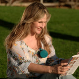 A woman sitting on a green lawn outdoors, smiling while reading a magazine. She is wearing a light floral blouse and light blue jeans, with a teal circular pendant necklace.