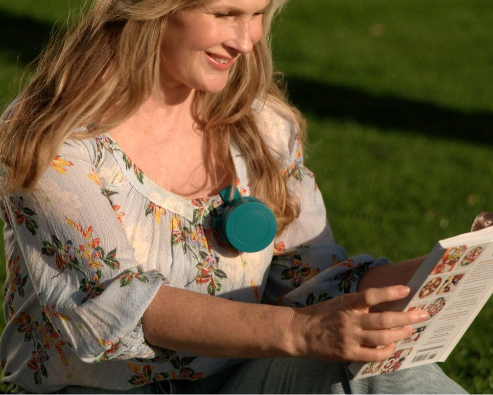 A woman sitting outdoors on grass, reading a booklet. She is wearing a light blue floral blouse and a teal fan is hooked to her shirt's neckline.