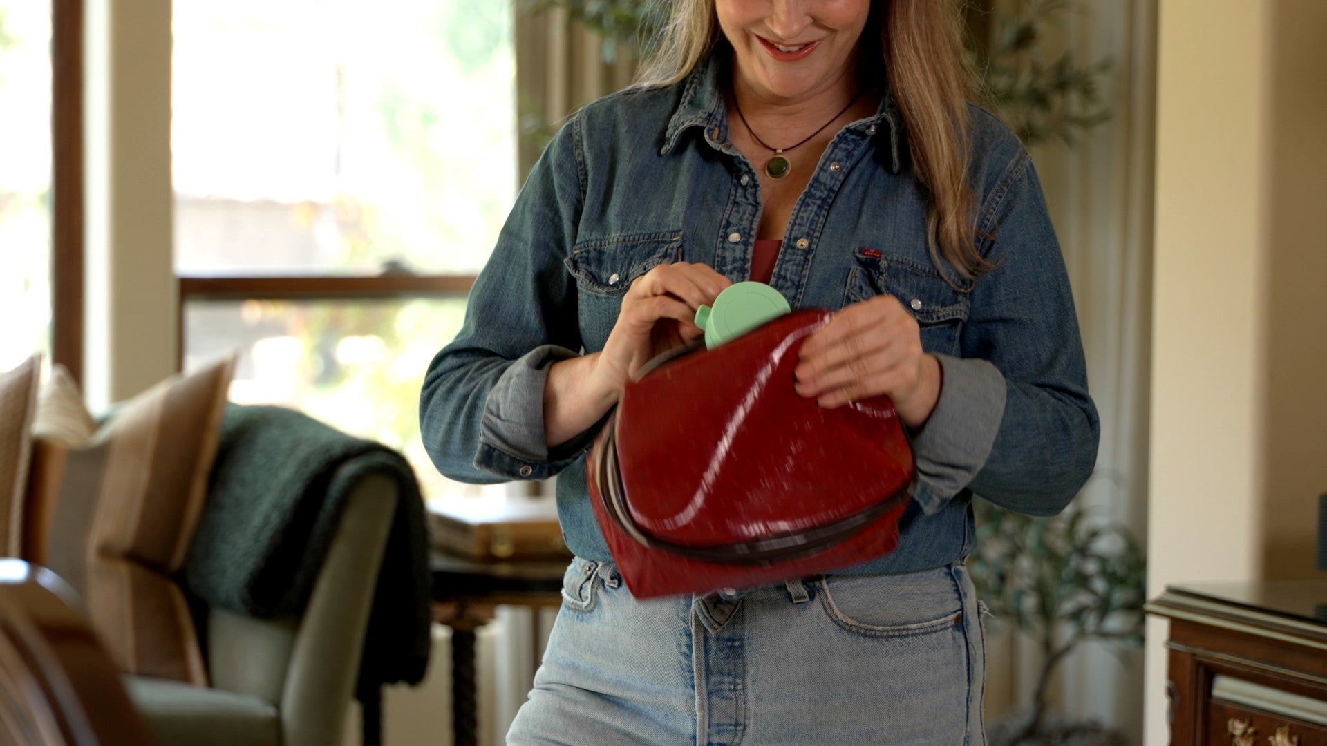 A woman in a denim shirt holding an open red leather cosmetic pouch, showing a light green compact fan inside, in a softly lit indoor setting.