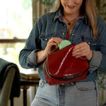 A woman in a denim shirt holding an open red leather cosmetic pouch, showing a light green compact fan inside, in a softly lit indoor setting.