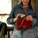 A woman in a denim shirt holding an open red leather cosmetic pouch, showing a light green compact fan inside, in a softly lit indoor setting.