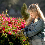A woman with blonde hair tied back, wearing a denim jacket and jeans, is gardening by a lake, touching a dense bush of bright pink flowers.