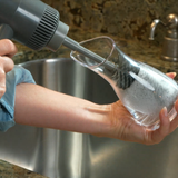 A person using a gray handheld air blower to blow air into a clear glass carafe sink top, set in a kitchen with a stainless steel sink and granite countertop.