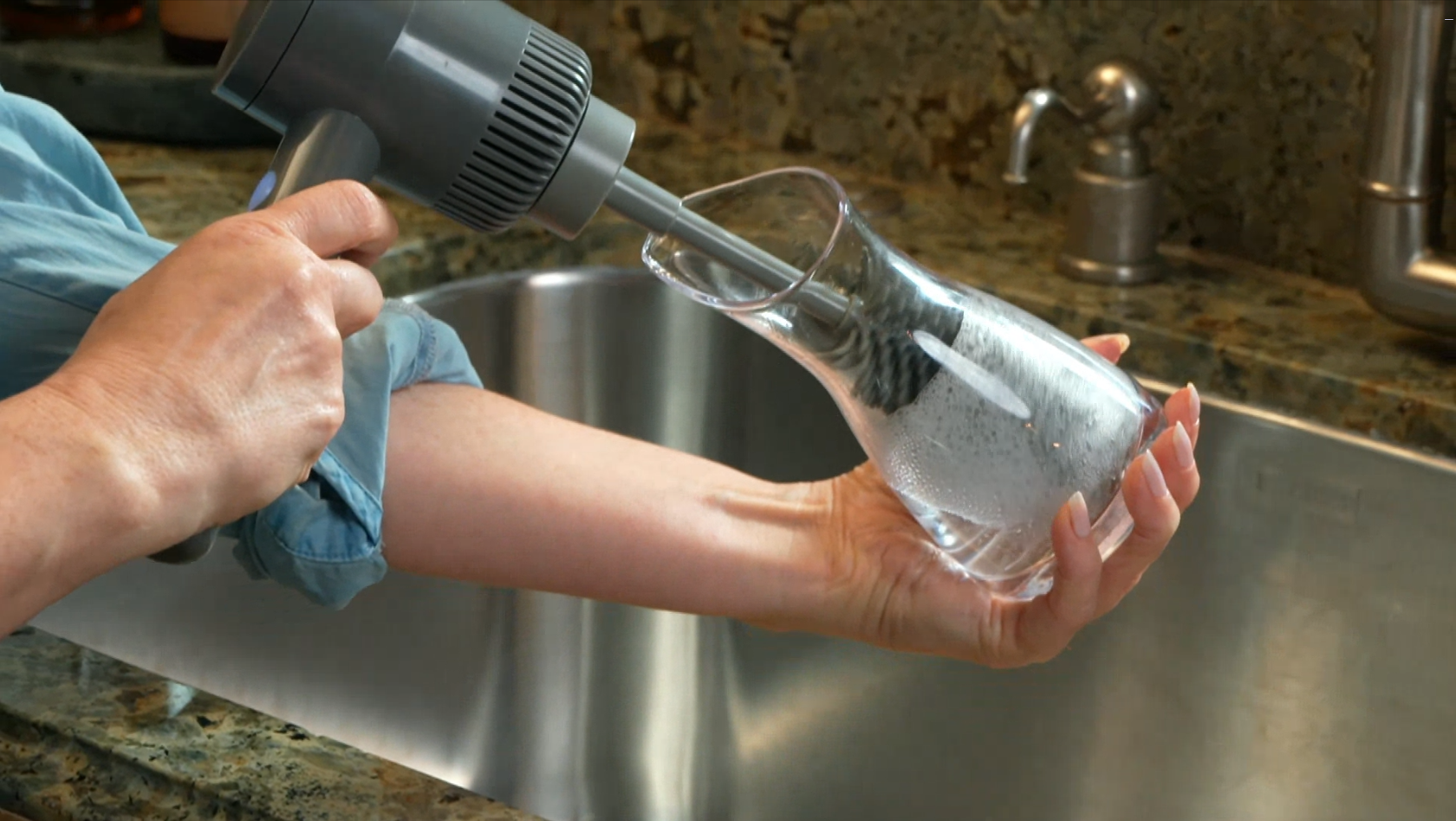 A person using a gray handheld air blower to blow air into a clear glass carafe sink top, set in a kitchen with a stainless steel sink and granite countertop.