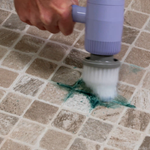 A close-up of a hand holding a lavender and gray handheld floor scrubber over a tiled surface, scrubbing a green stain into the grout lines.