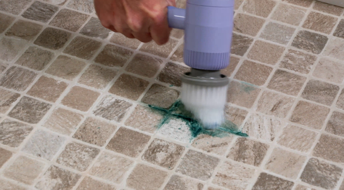 A close-up of a hand holding a lavender and gray handheld floor scrubber over a tiled surface, scrubbing a green stain into the grout lines.